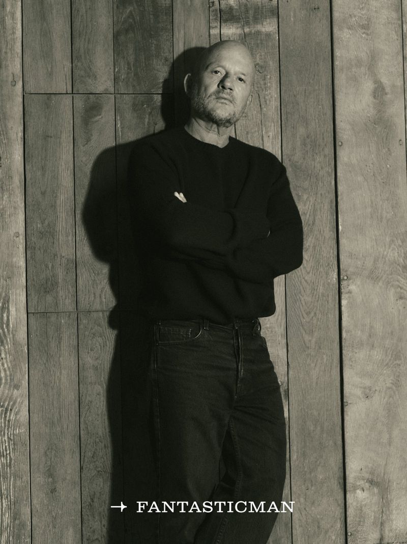 Portrait of architect Vincent Van Duysen standing against a vertical wood plank wall wearing a dark sweater and jeans