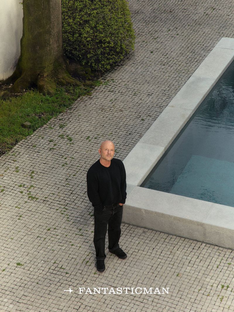Aerial portrait of bald man in black sweater standing beside a rectangular swimming pool on cobblestone patio