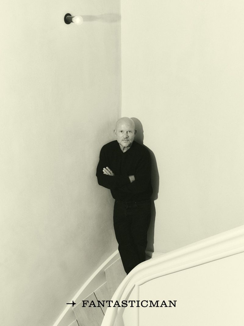 A bald man in a black outfit stands on stairs next to a white wall with a light fixture. He is featured in a magazine ad.