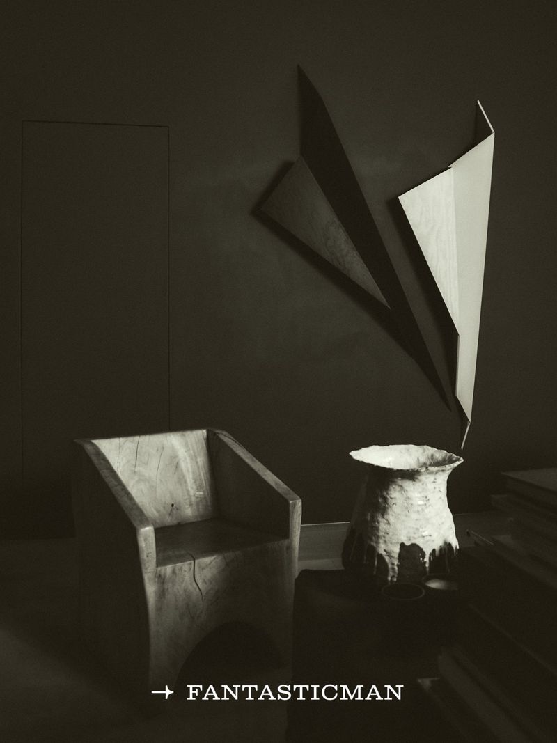 Minimalist interior with a carved wooden chair, a textured ceramic vessel, and geometric wall sculptures under dark lighting.