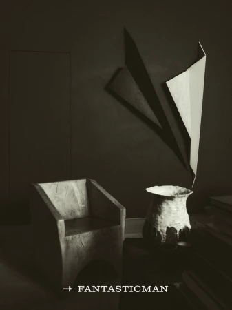 Minimalist interior with a carved wooden chair, a textured ceramic vessel, and geometric wall sculptures under dark lighting.
