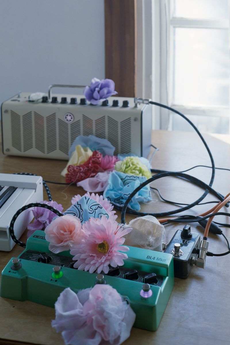 Musical equipment including a green guitar pedal and beige amplifier decorated with colorful artificial flowers on a wooden table.
