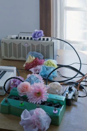 Musical equipment including a green guitar pedal and beige amplifier decorated with colorful artificial flowers on a wooden table.