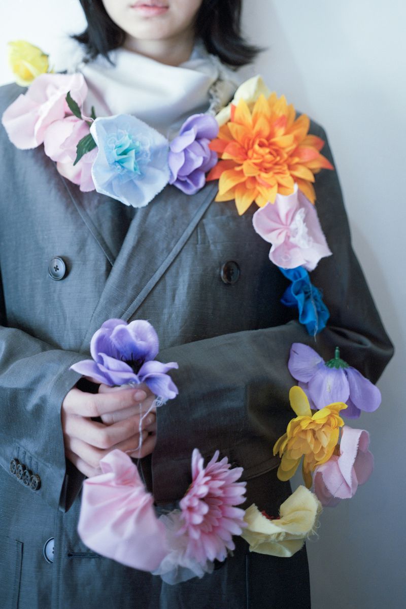 Model wearing a dark grey coat adorned with a vibrant floral garland for The Plant Magazine editorial Symphony in Bloom