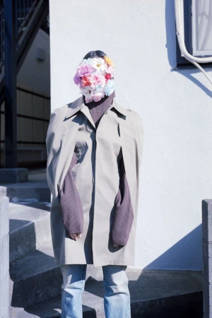 Model wearing a beige coat and floral face covering standing outdoors against a white wall.
