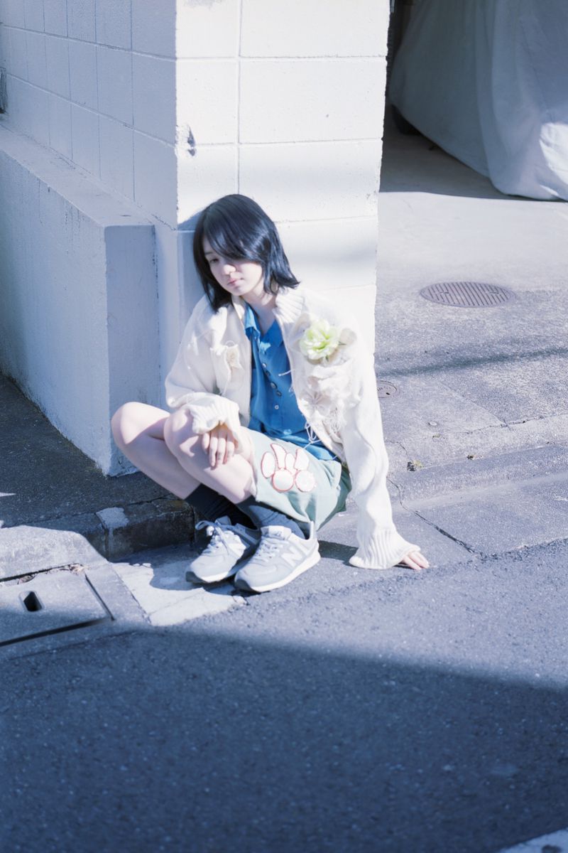 Young model with dark hair wearing a cream cardigan, blue shirt, and grey sneakers crouching against a white tiled wall.