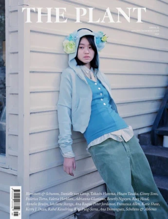 Young woman in a light blue hooded sweatshirt and green trousers leaning against a white clapboard wall
