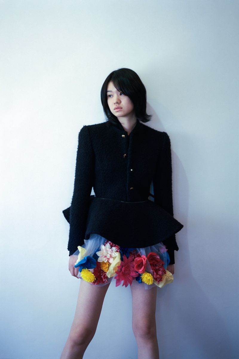 Model wearing a black jacket and floral-trimmed tulle skirt against a white wall.