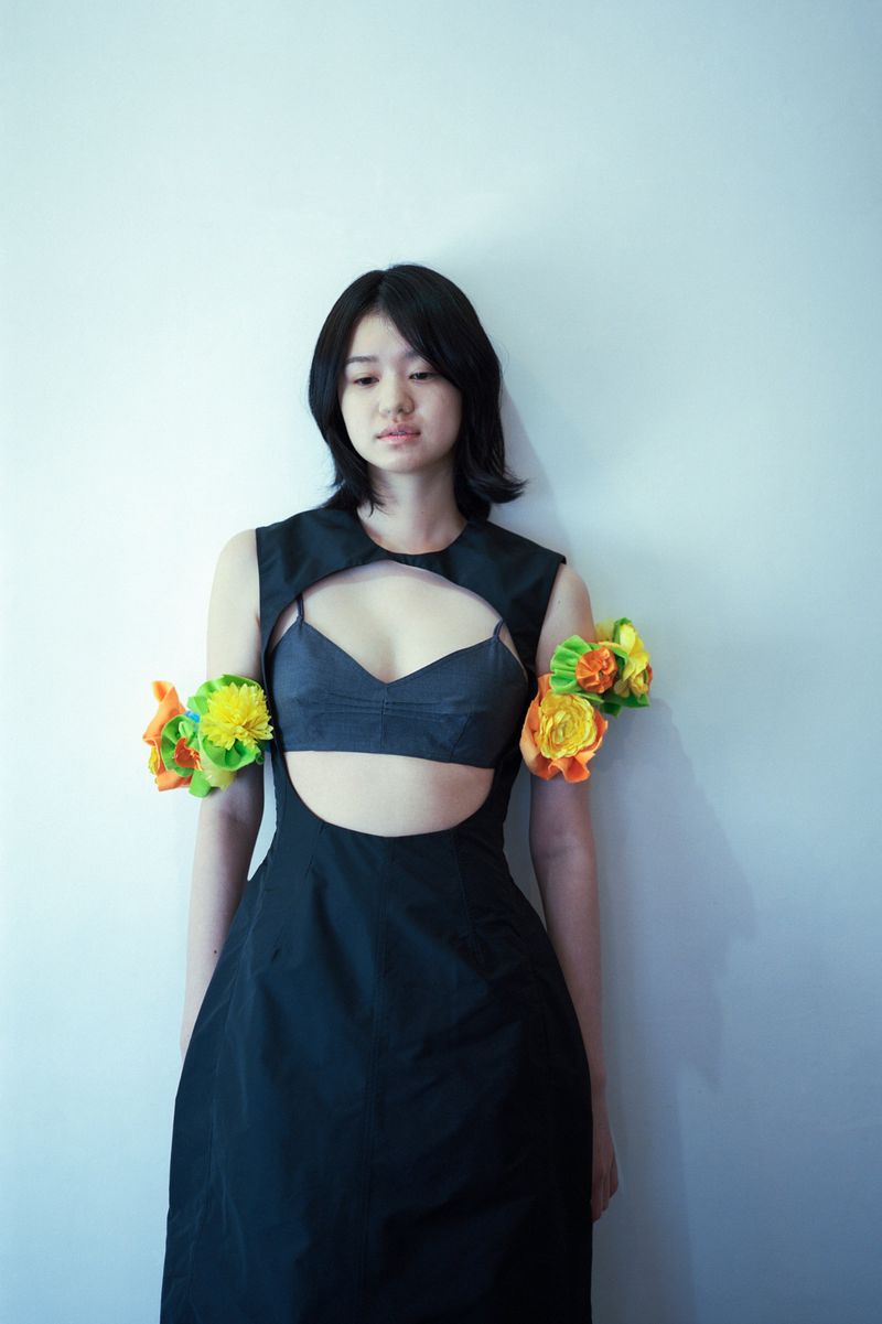 Young woman in a black cutout dress adorned with bright yellow and orange floral armbands against a plain white wall