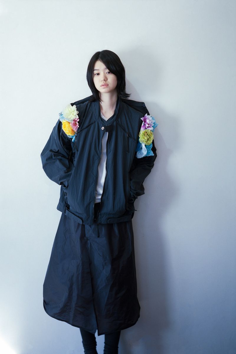 Portrait of a stylish Asian woman wearing a dark blue jacket with colorful flower accents, posing against a light gray wall indoors.