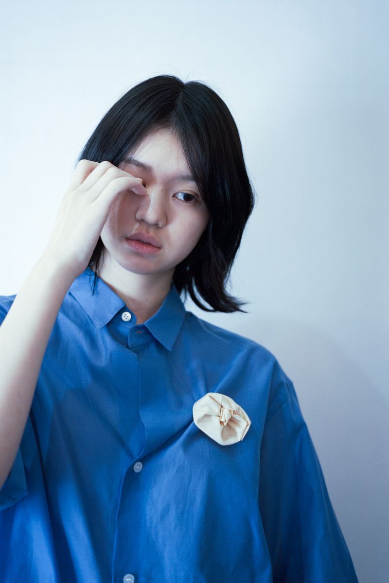 Young woman in a vibrant blue collared shirt touching her eye against a plain white wall background