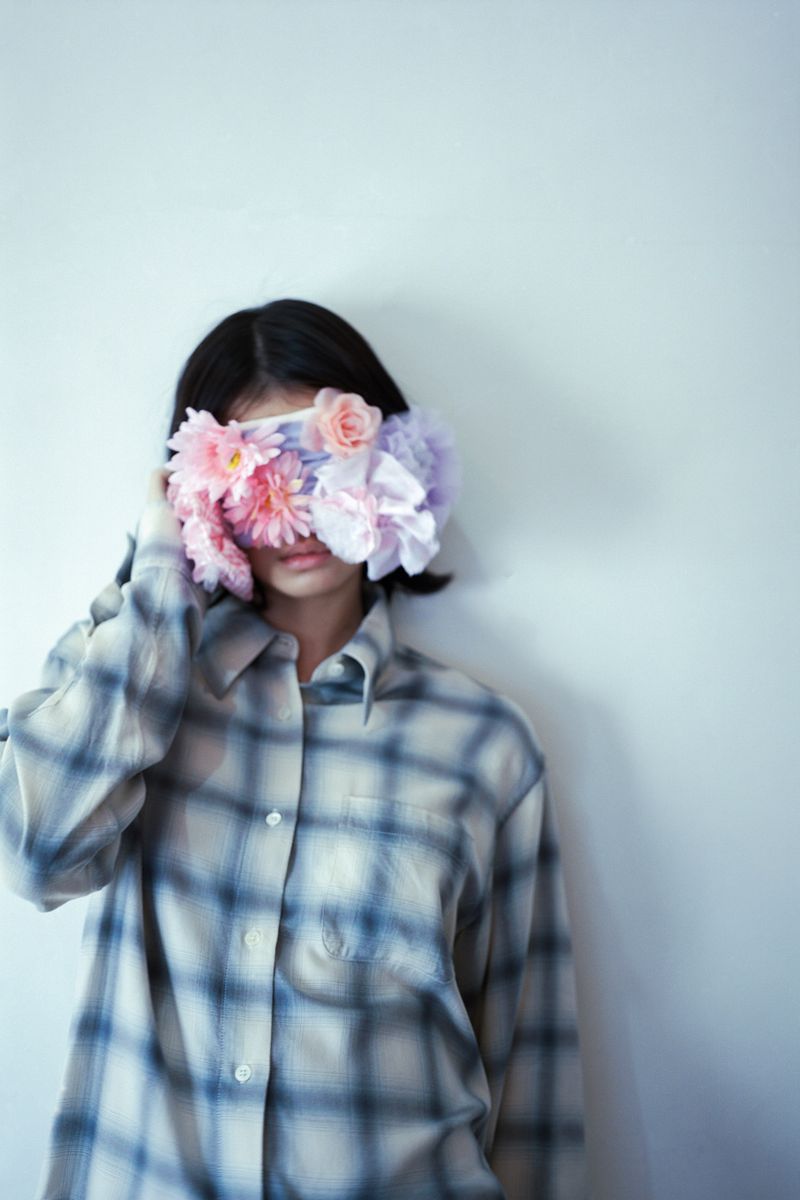 Woman wearing a plaid shirt with floral arrangements covering her eyes against a light background