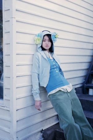 Young woman wearing a hooded sweatshirt adorned with floral headpieces leaning against white wooden siding for an editorial.
