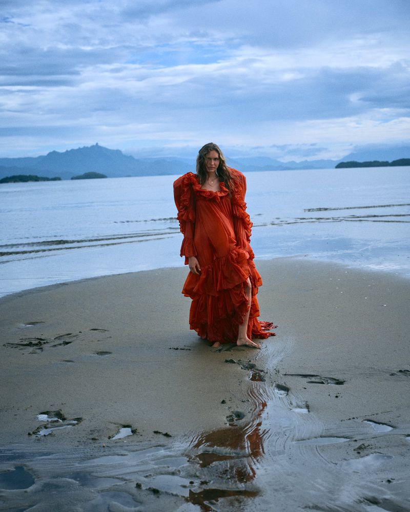 Model Angelina Kendall in a red ruffled gown standing on a sandy shore by water under an overcast sky for Financial Times HTSI Magazine