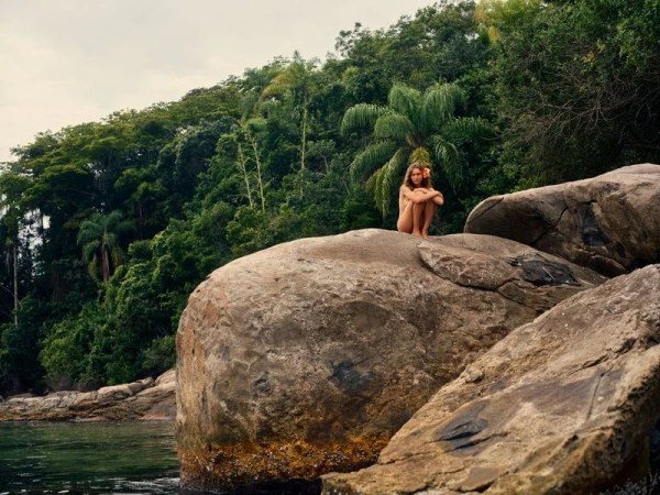 Model Angelina Kendall crouched on a large coastal rock surrounded by dense tropical forest for HTSI Magazine.