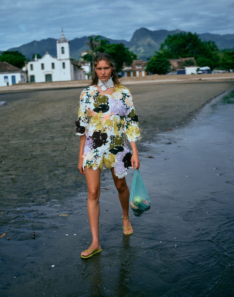 Model walking on a sandy beach holding a green plastic bag with a town and mountains in the background