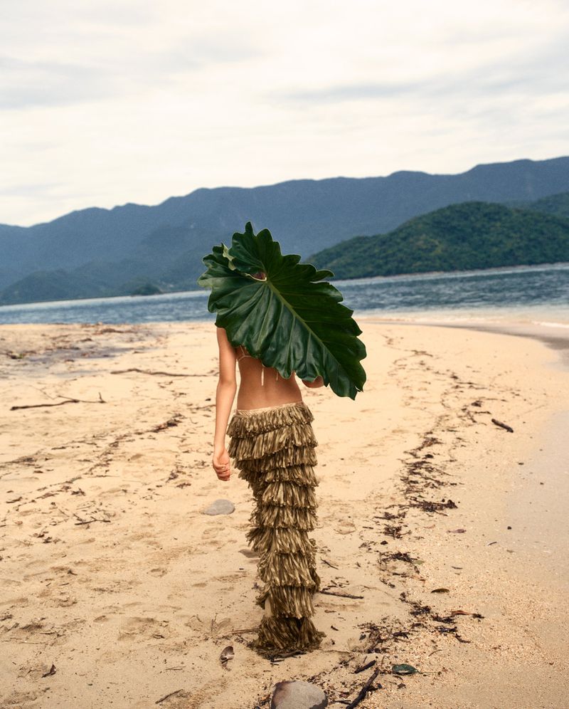 Model Angelina Kendall holding a large green leaf on a sandy beach for Tropic Of Venus editorial by Josh Olins