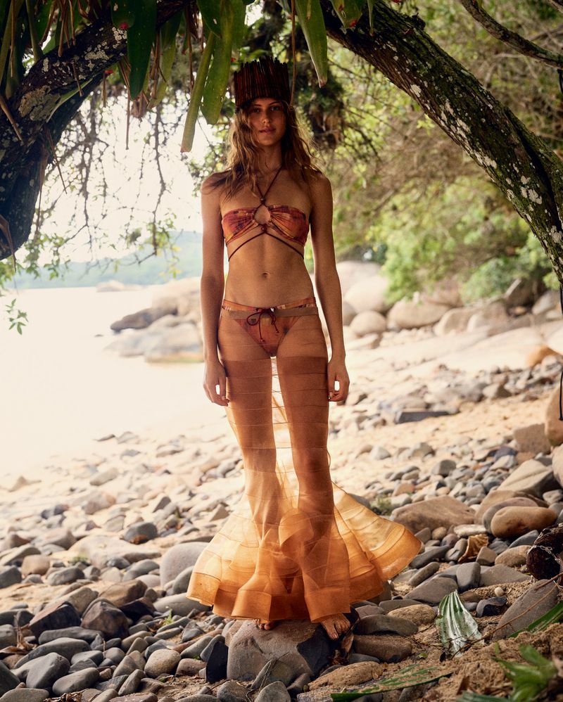 Model Angelina Kendall in a patterned bikini and sheer tiered skirt on a sandy beach under tree branches for HTSI Magazine.