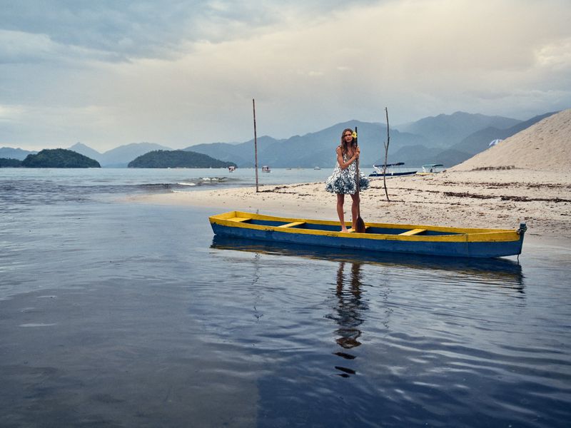 Model standing on a blue and yellow canoe in calm water near a sandy beach with mountains in the background
