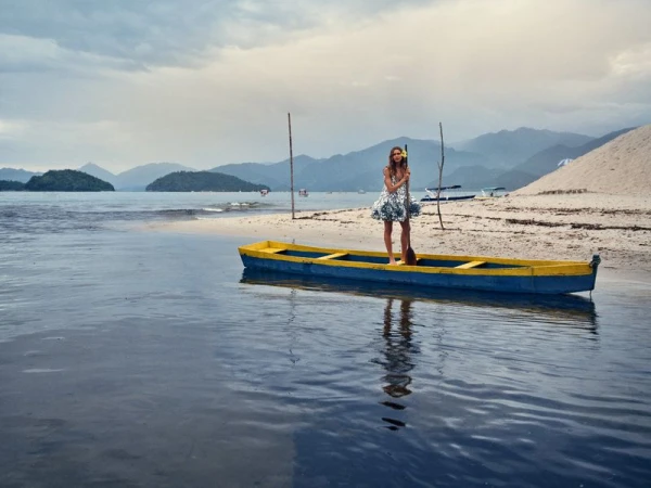 Model standing on a blue and yellow canoe in calm water near a sandy beach with mountains in the background