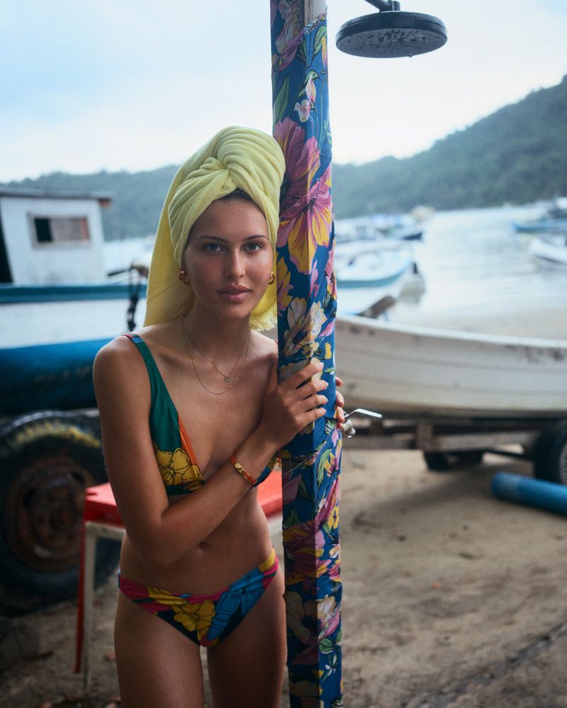 Model Angelina Kendall in a floral bikini with a yellow towel on her head standing by a beach shower for Financial Times HTSI Magazine.