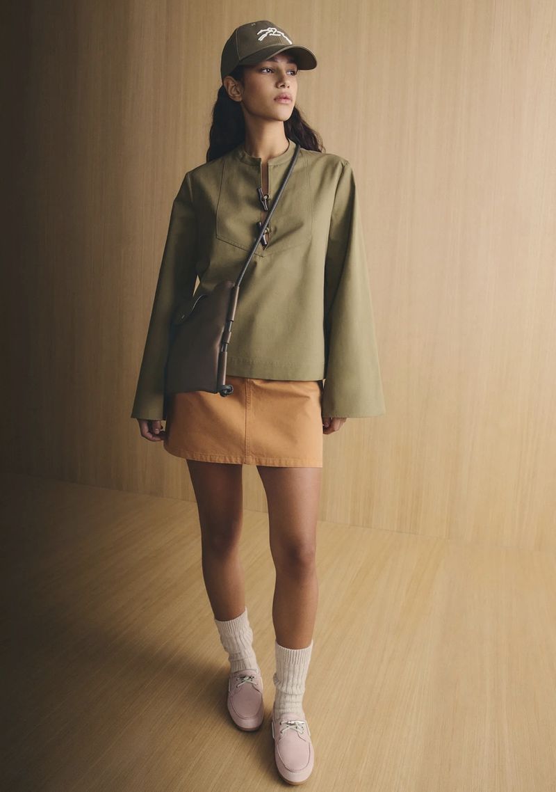 Model wearing a green top, orange skirt, baseball cap, and crossbody bag against a warm wood-paneled background.