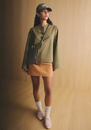 Model wearing a green top, orange skirt, baseball cap, and crossbody bag against a warm wood-paneled background.