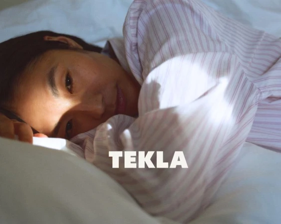 Asian woman in pink-striped pajama top lies on white bedding with a tranquil expression and soft lighting; sleepwear fashion shot.