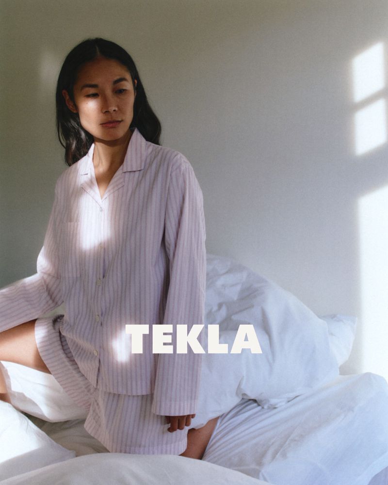 Woman in pink striped pajama set kneeling on a white bed with sunlight streaming across the wall.
