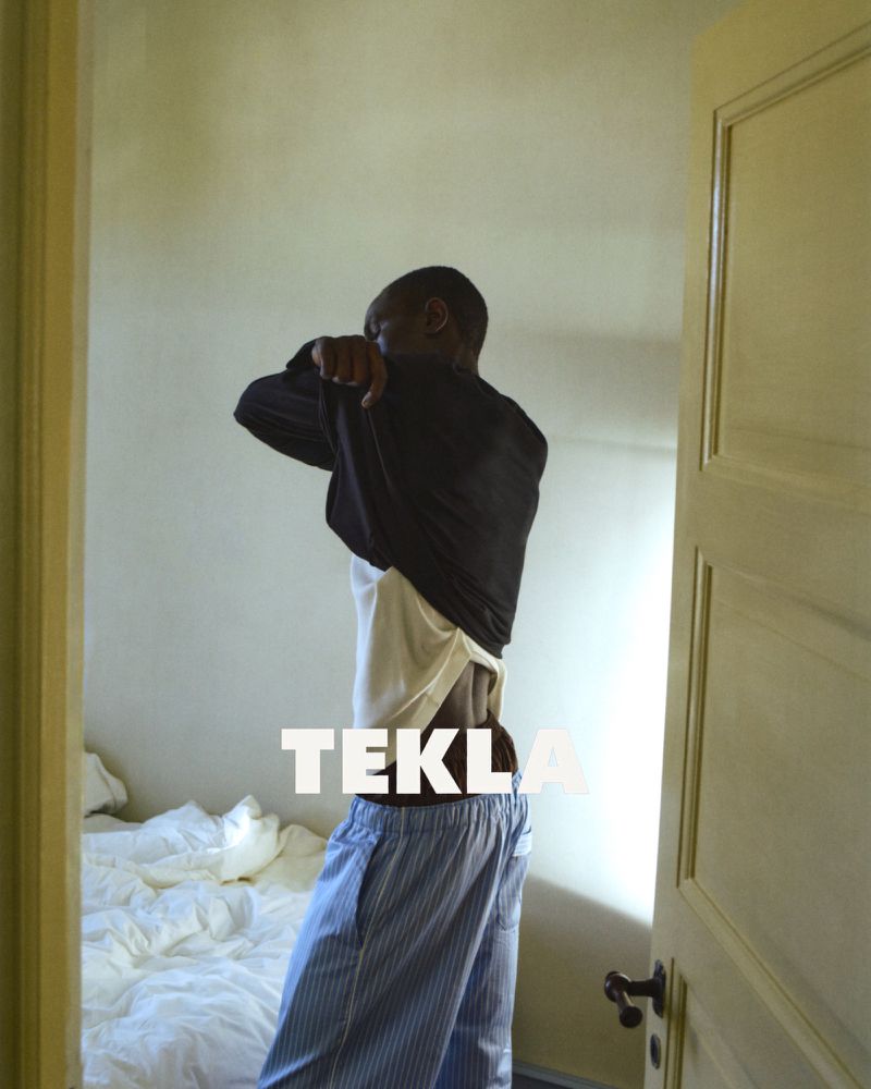 A Black man taking off his black shirt while wearing blue striped pajama pants in a bedroom for Tekla Sleepwear.