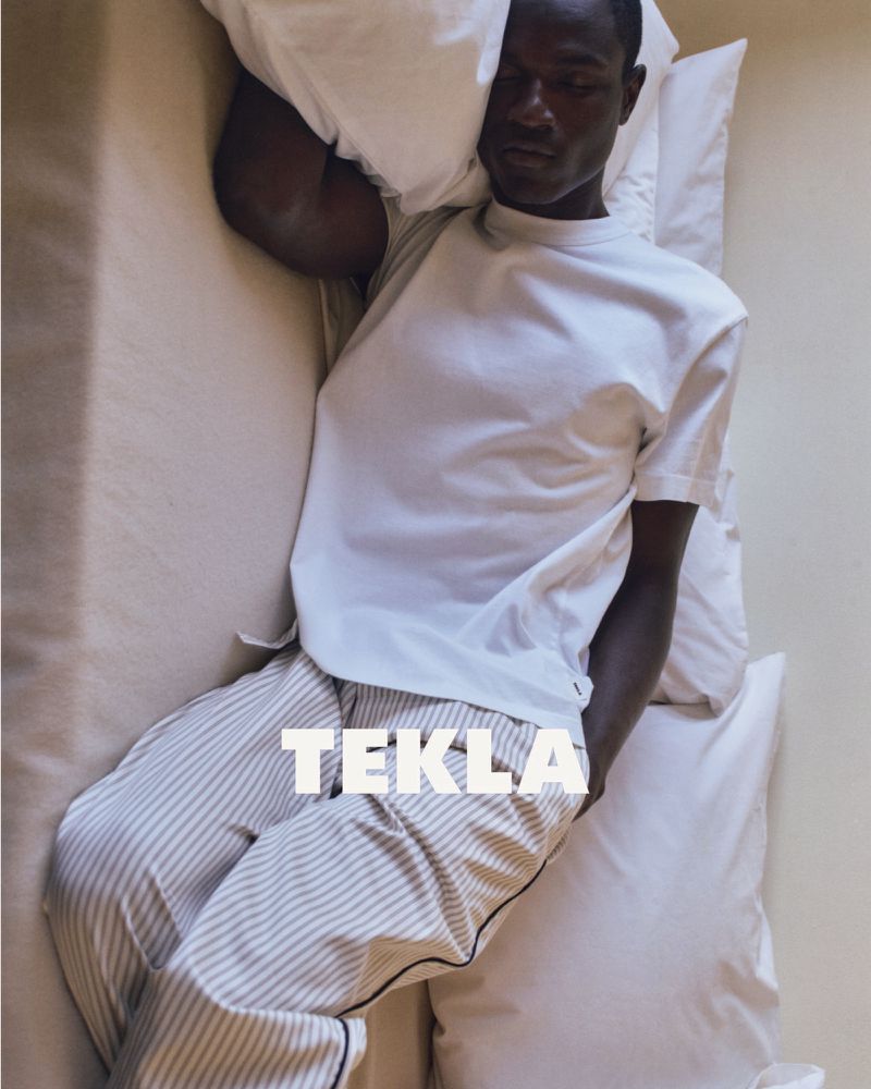 Black man lying in bed wearing a white t-shirt and striped pyjama trousers, resting against white pillows.