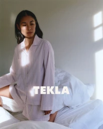 Woman in pink striped pajama set kneeling on a white bed with sunlight streaming across the wall.