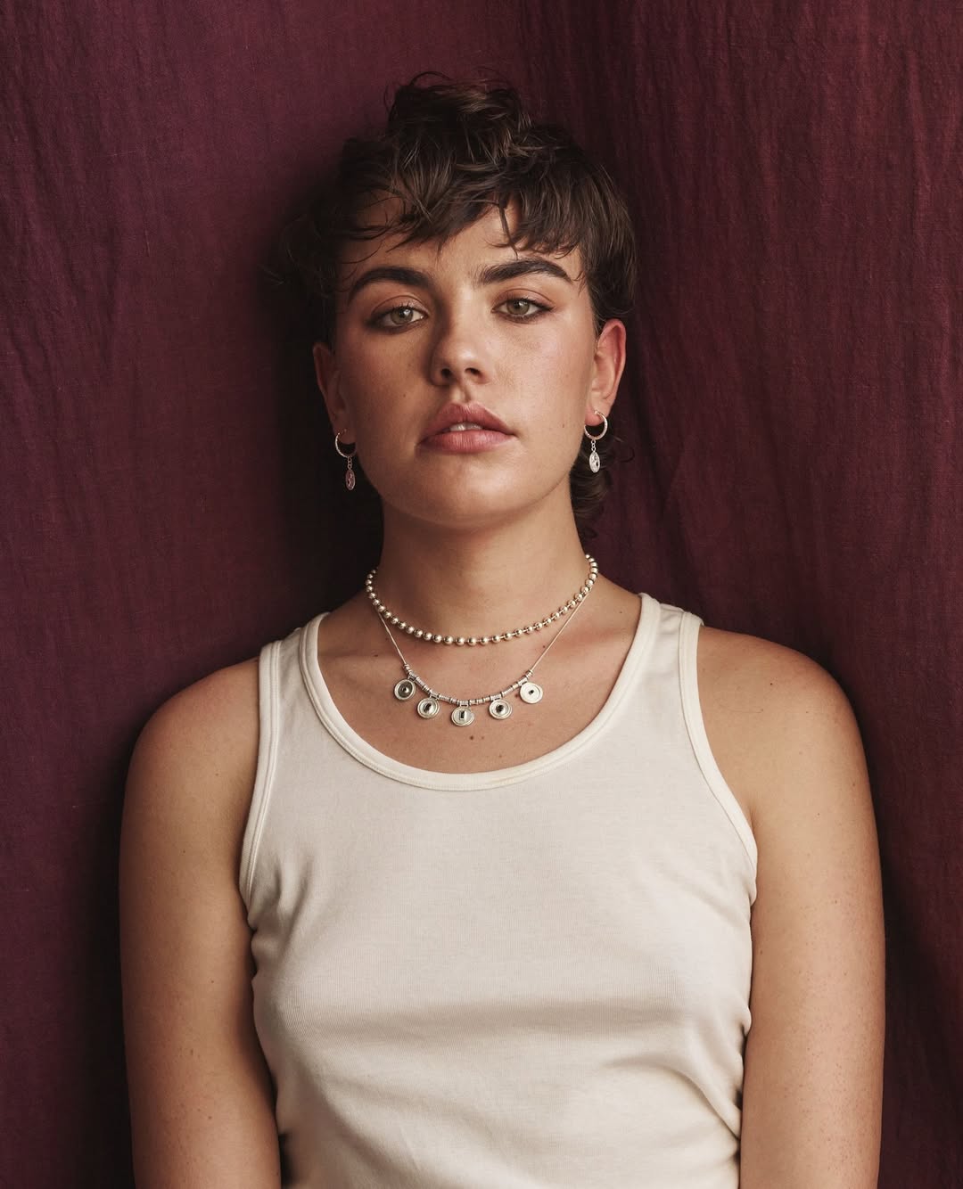 Close-up studio portrait of a young woman with short dark hair wearing a white tank top and layered necklaces in front of a maroon backdrop.