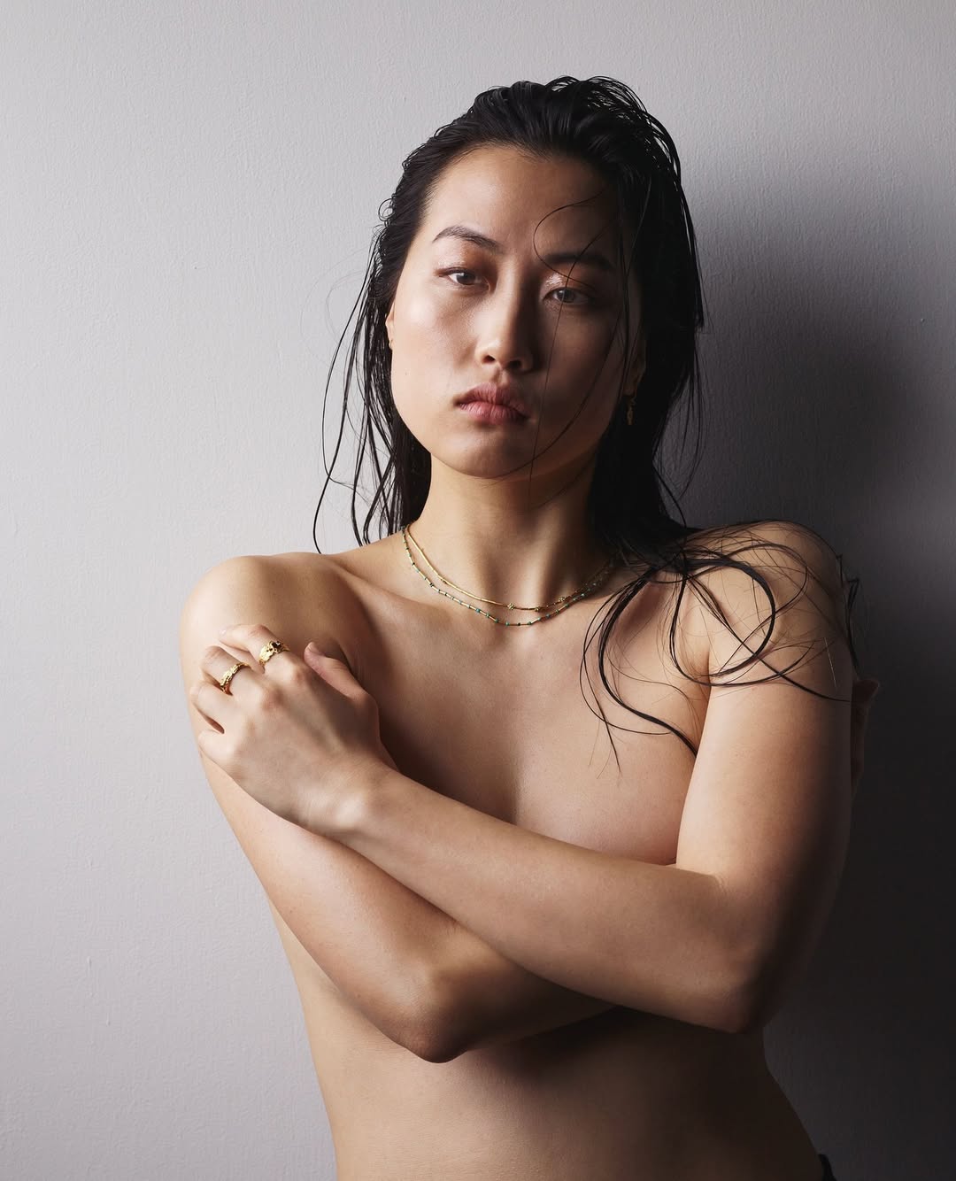 Headshot of an Asian woman with wet dark hair, gold necklace, and ring against a neutral gray background in a portrait style.