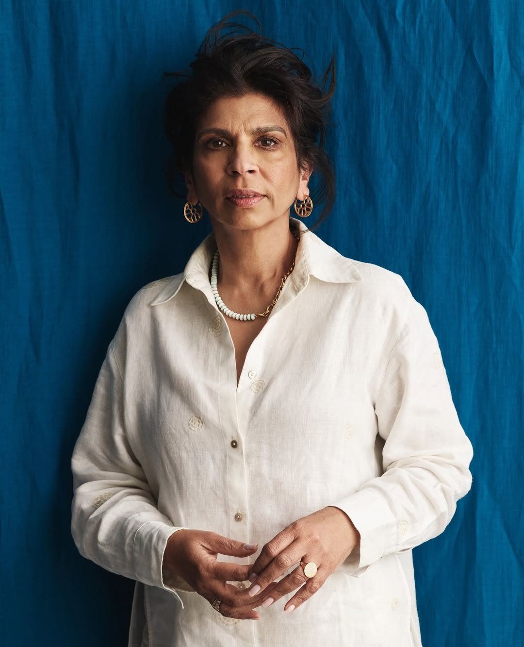 Portrait of a confident woman in a white shirt against a blue backdrop. She is wearing jewelry, has short dark hair, and is facing forward.