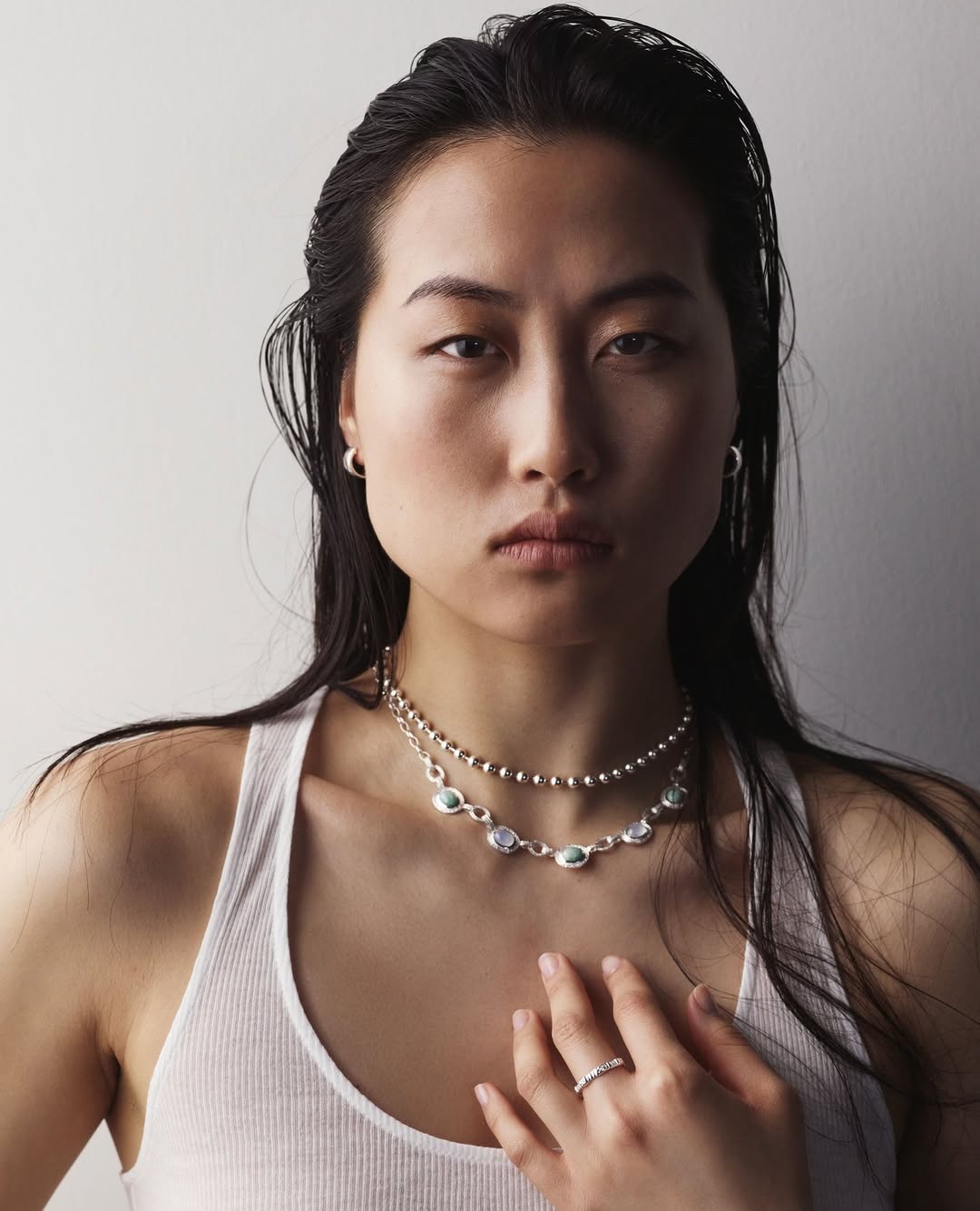 Close-up of an Asian woman with dark hair, wearing jewelry and a white tank top, posing in front of a plain light gray backdrop.