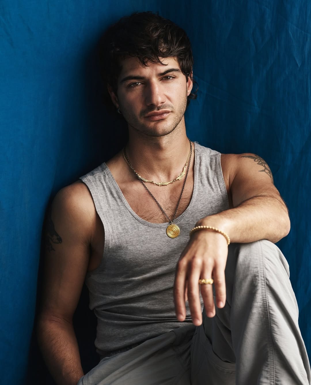 Portrait of a young adult man with dark hair, wearing a gray tank top and gold jewelry, against a textured blue background.