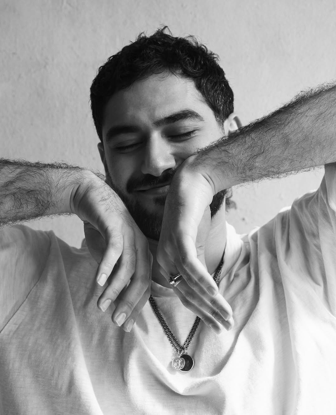 Black and white portrait of a man with dark hair and a beard, his hands near his face, wearing a t-shirt and chain necklace indoors.