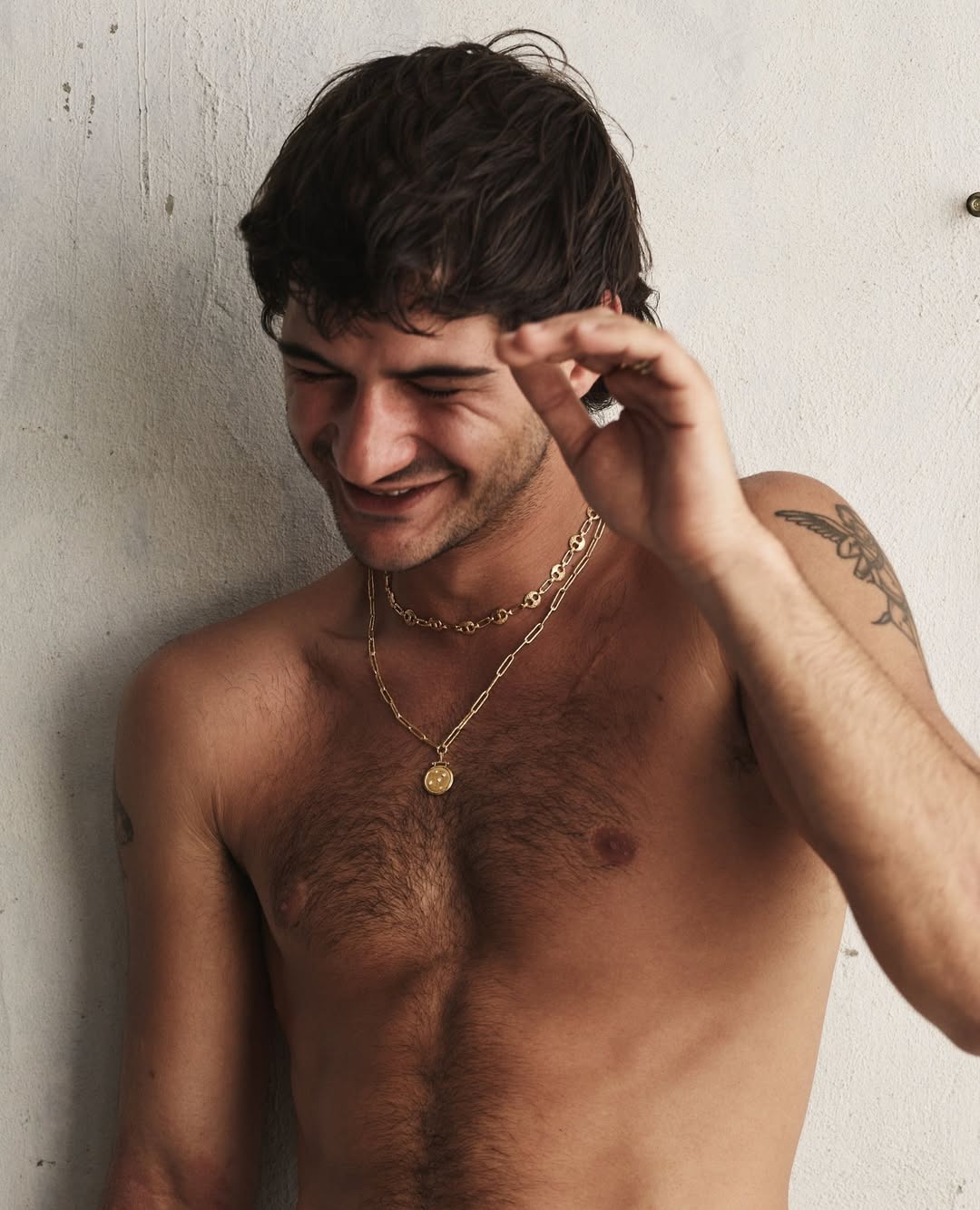 Man with dark hair smiling, wearing two gold chains against a textured wall, portrait shot.