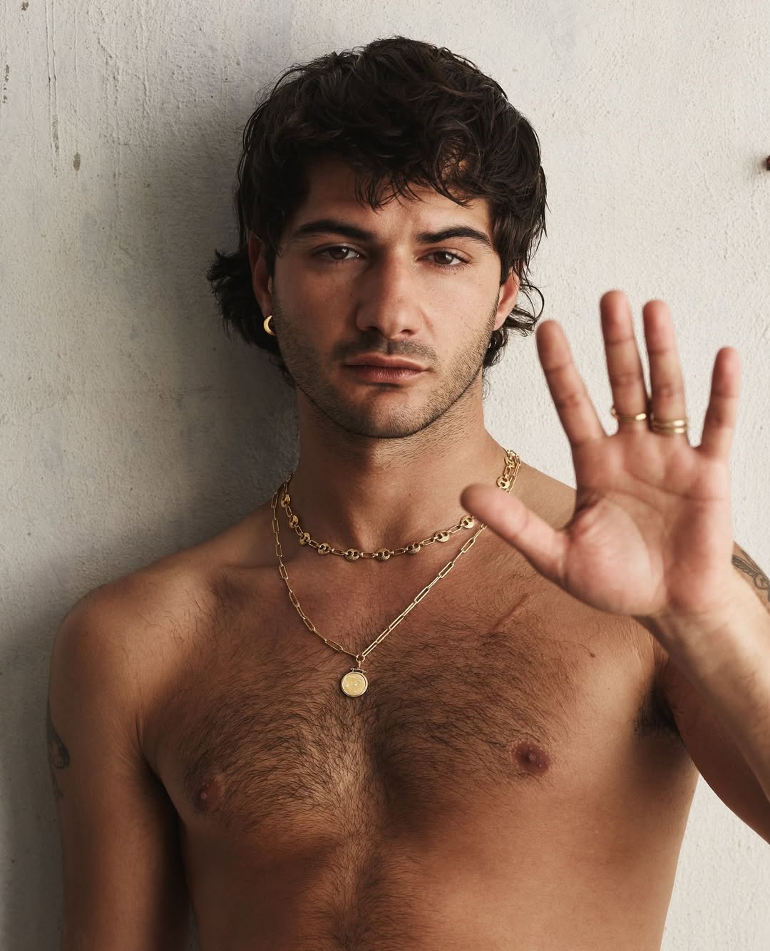 A young adult male with dark hair, wearing gold jewelry, poses against a neutral wall. He has his hand up, showing several rings.