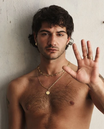 A young adult male with dark hair, wearing gold jewelry, poses against a neutral wall. He has his hand up, showing several rings.