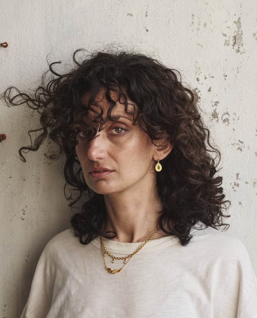 Portrait of a woman with curly hair wearing gold jewelry against a textured wall. Close-up shot featuring fashion and beauty elements.