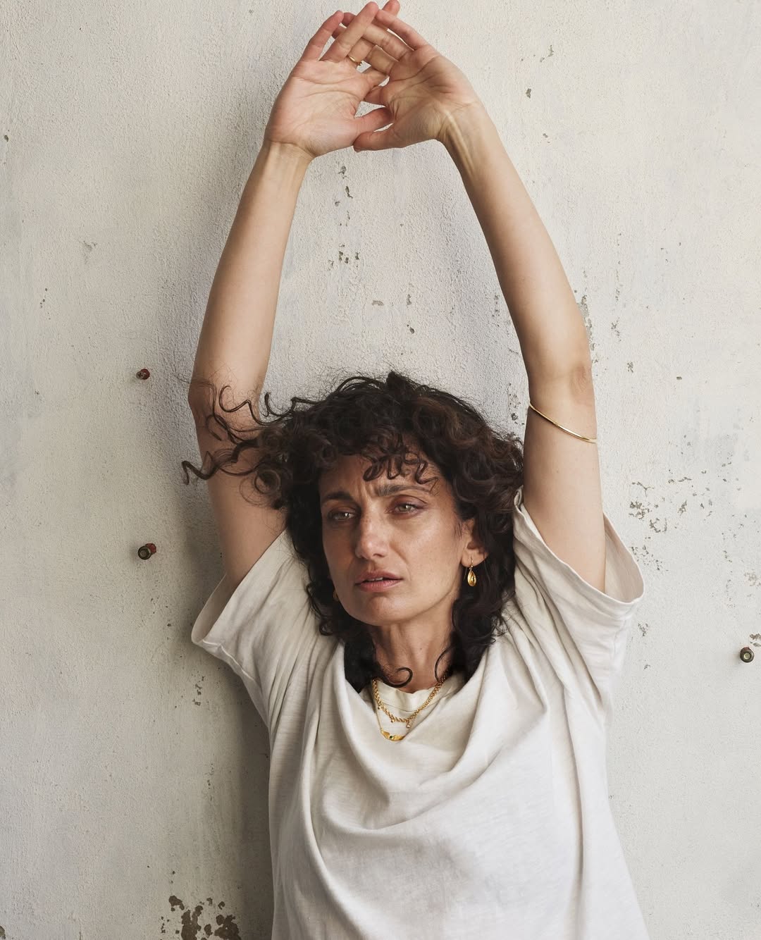 Portrait of a woman with curly brown hair and a neutral expression. She is wearing a white t-shirt and gold jewelry with her arms raised overhead.