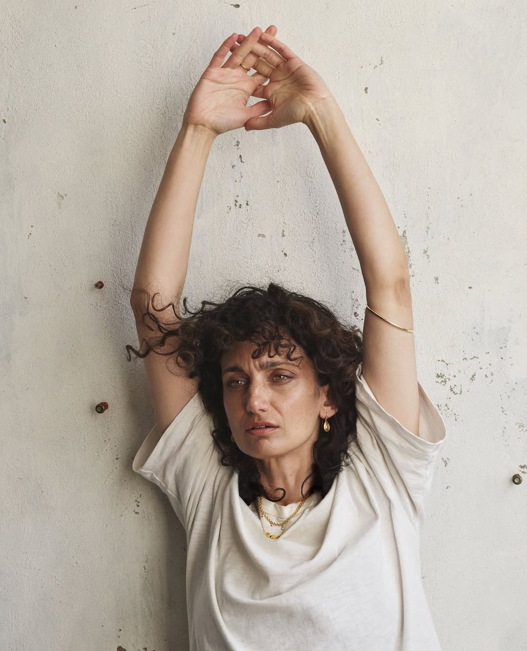 Portrait of a woman with curly hair wearing a white t-shirt, arms raised, against a textured neutral wall indoors.