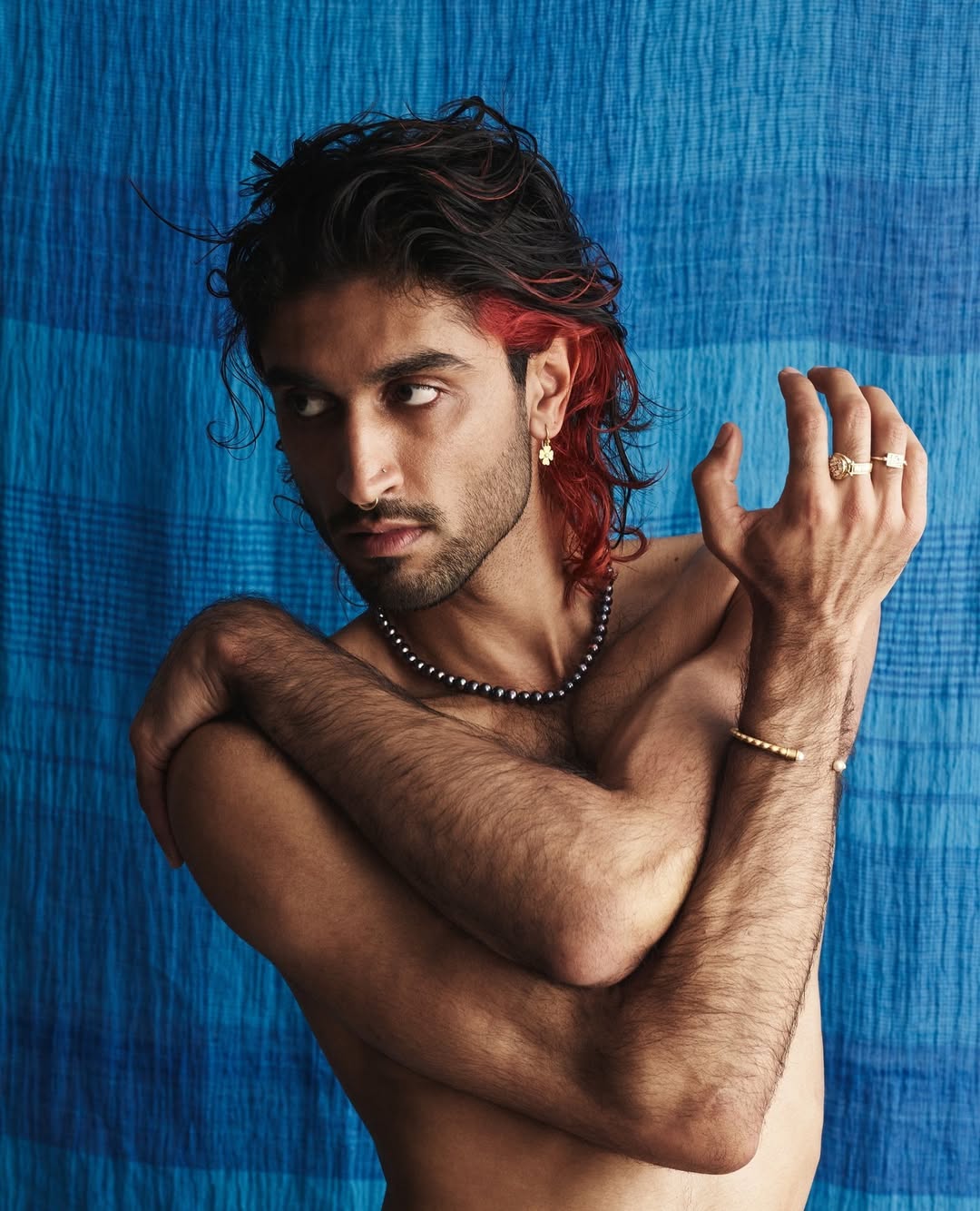 Portrait of a bare-chested man with dyed red hair posing against a blue patterned backdrop, wearing pearl necklace, gold rings and bracelet.