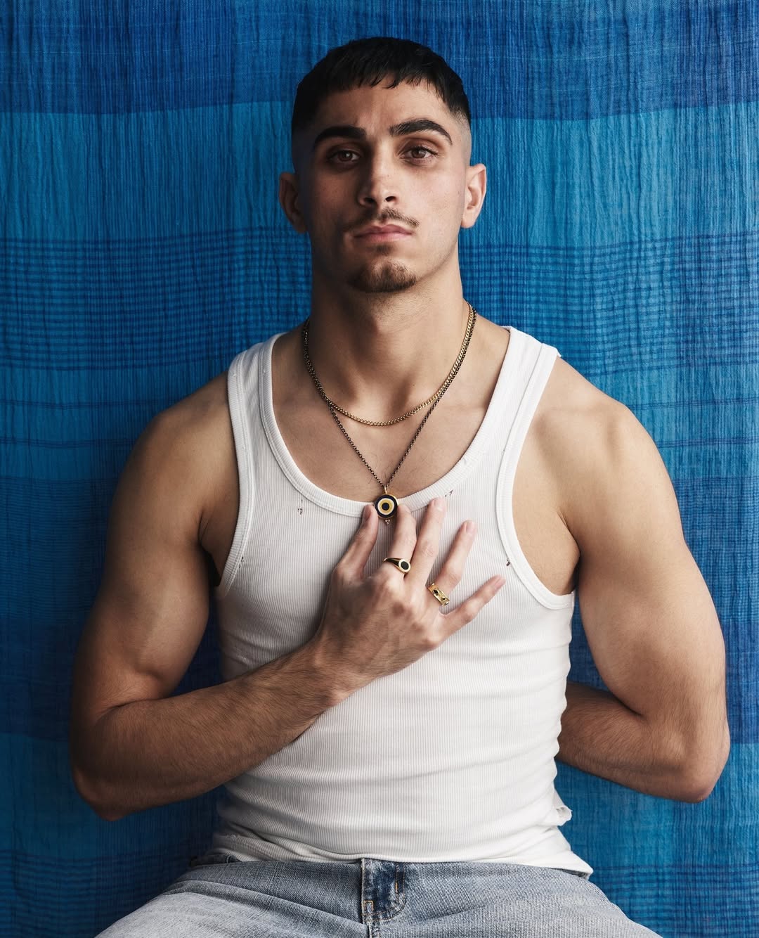 Portrait of a young adult male with dark hair and light blue jeans, wearing a white tank top against a striped blue background.