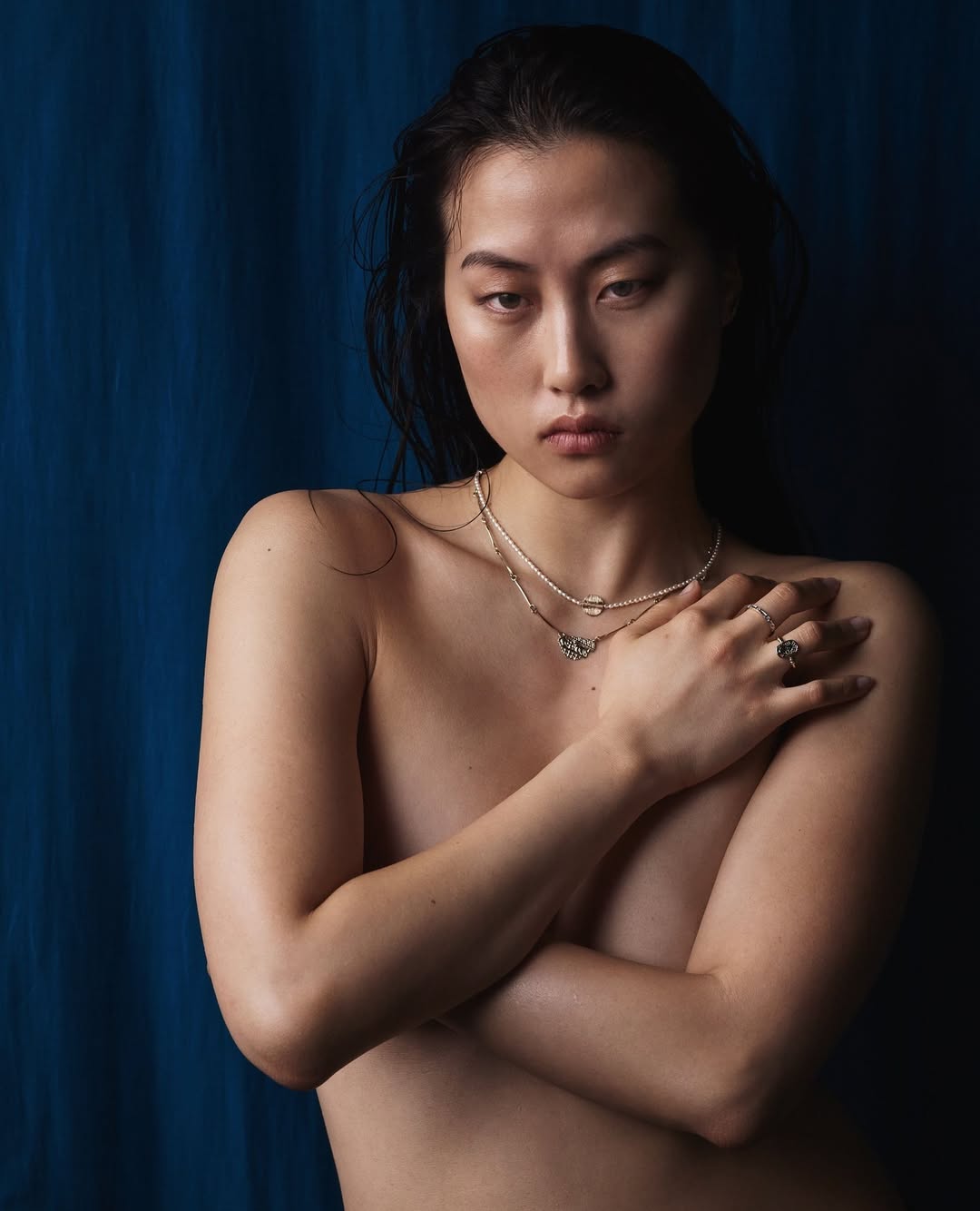 Striking portrait of an Asian adult female with wet hair and neutral expression, adorned with jewelry, against a deep blue background.