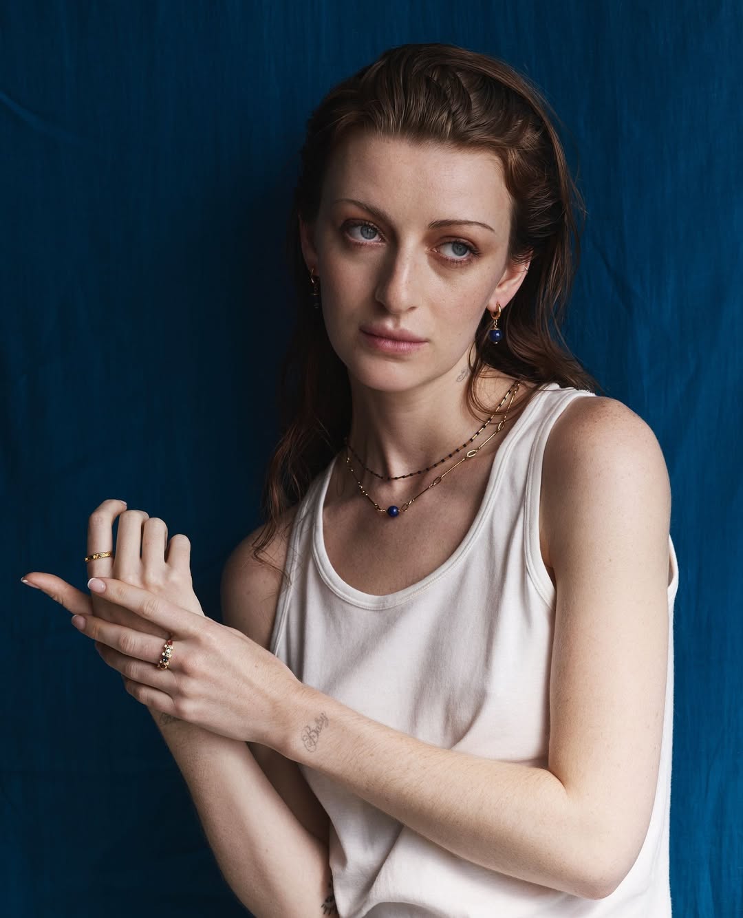 Close-up of a woman with red hair wearing a white tank top and jewelry, posing against a dark blue background, showcasing beauty.