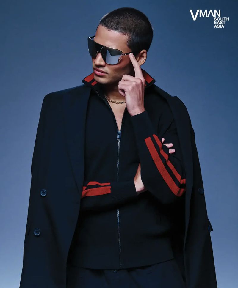 Portrait of a handsome male model wearing sunglasses, a black jacket with red stripes, and a gold chain against a gradient blue backdrop.