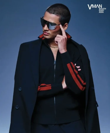 Portrait of a handsome male model wearing sunglasses, a black jacket with red stripes, and a gold chain against a gradient blue backdrop.